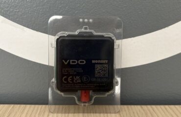 Antena DSRC i moduli Meter navishcho vony potribni pry perevirtsi takhohrafiv Black square electronic module labeled 'VDO NORBIT' in a clear protective case, with a red connector at the bottom, placed on a wood table against a gray wall with white curved stripes