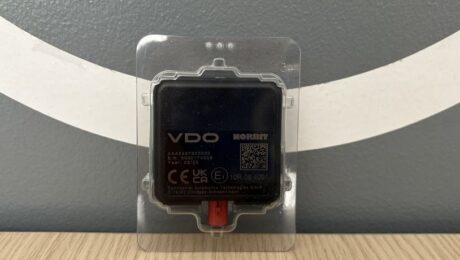Antena DSRC i moduli Meter navishcho vony potribni pry perevirtsi takhohrafiv Black square electronic module labeled 'VDO NORBIT' in a clear protective case, with a red connector at the bottom, placed on a wood table against a gray wall with white curved stripes