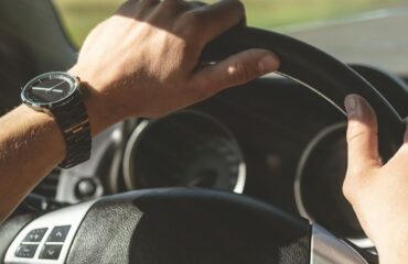 Driver's hands on a car steering wheel, wearing a metal wristwatch on the left wrist.