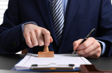 положення Business professional in a navy suit stamping a document with a wooden stamp, pen in the other hand on a desk.