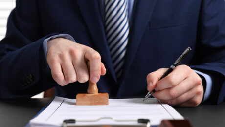 положення Business professional in a navy suit stamping a document with a wooden stamp, pen in the other hand on a desk.