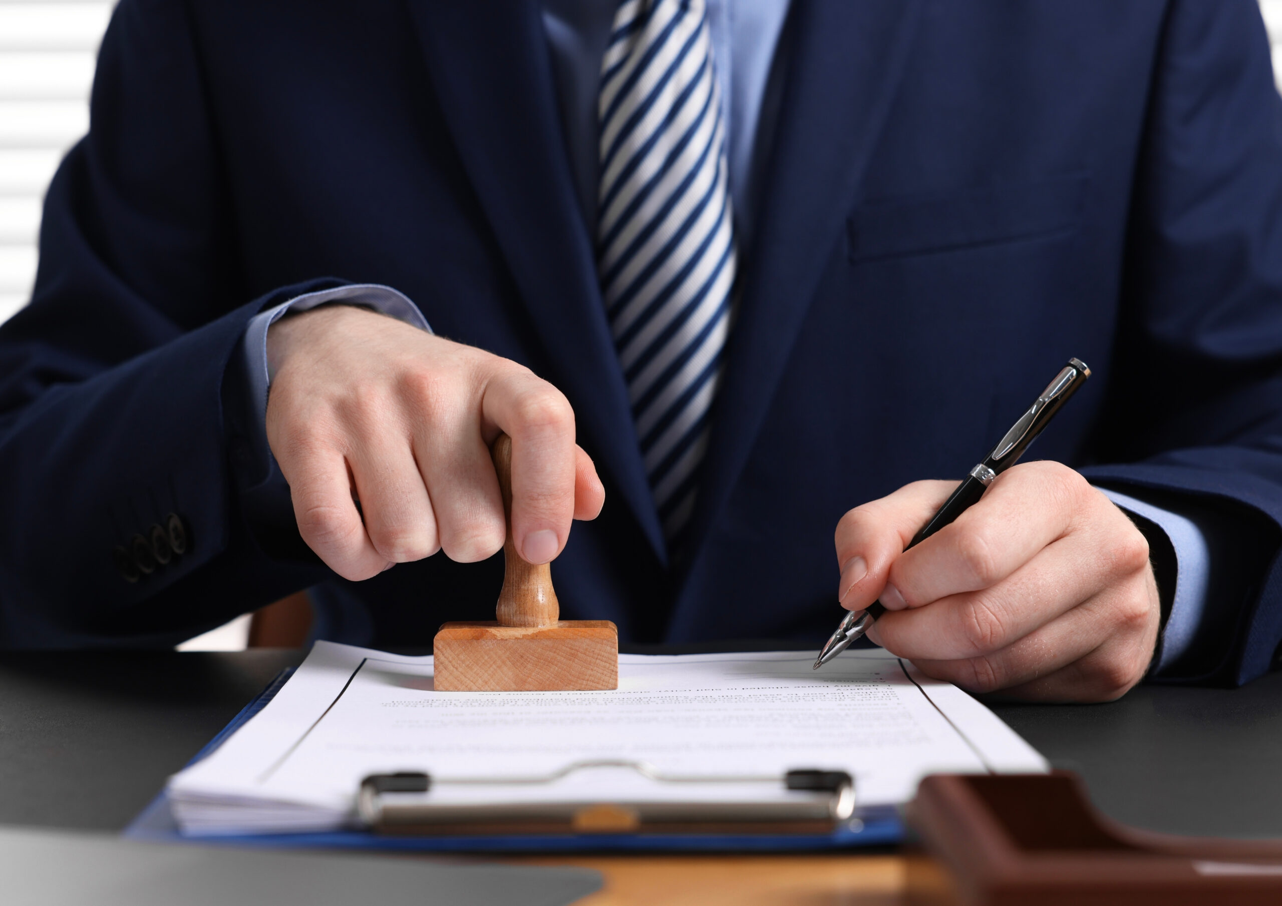 положення Business professional in a navy suit stamping a document with a wooden stamp, pen in the other hand on a desk.