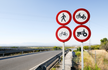 пдр Cluster of four red-and-white no-entry traffic signs: pedestrian, bicycle, motorcycle, and tractor rider on a roadside backdrop with blue sky and desert landscape.