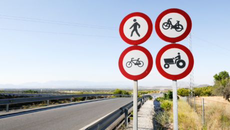 пдр Cluster of four red-and-white no-entry traffic signs: pedestrian, bicycle, motorcycle, and tractor rider on a roadside backdrop with blue sky and desert landscape.