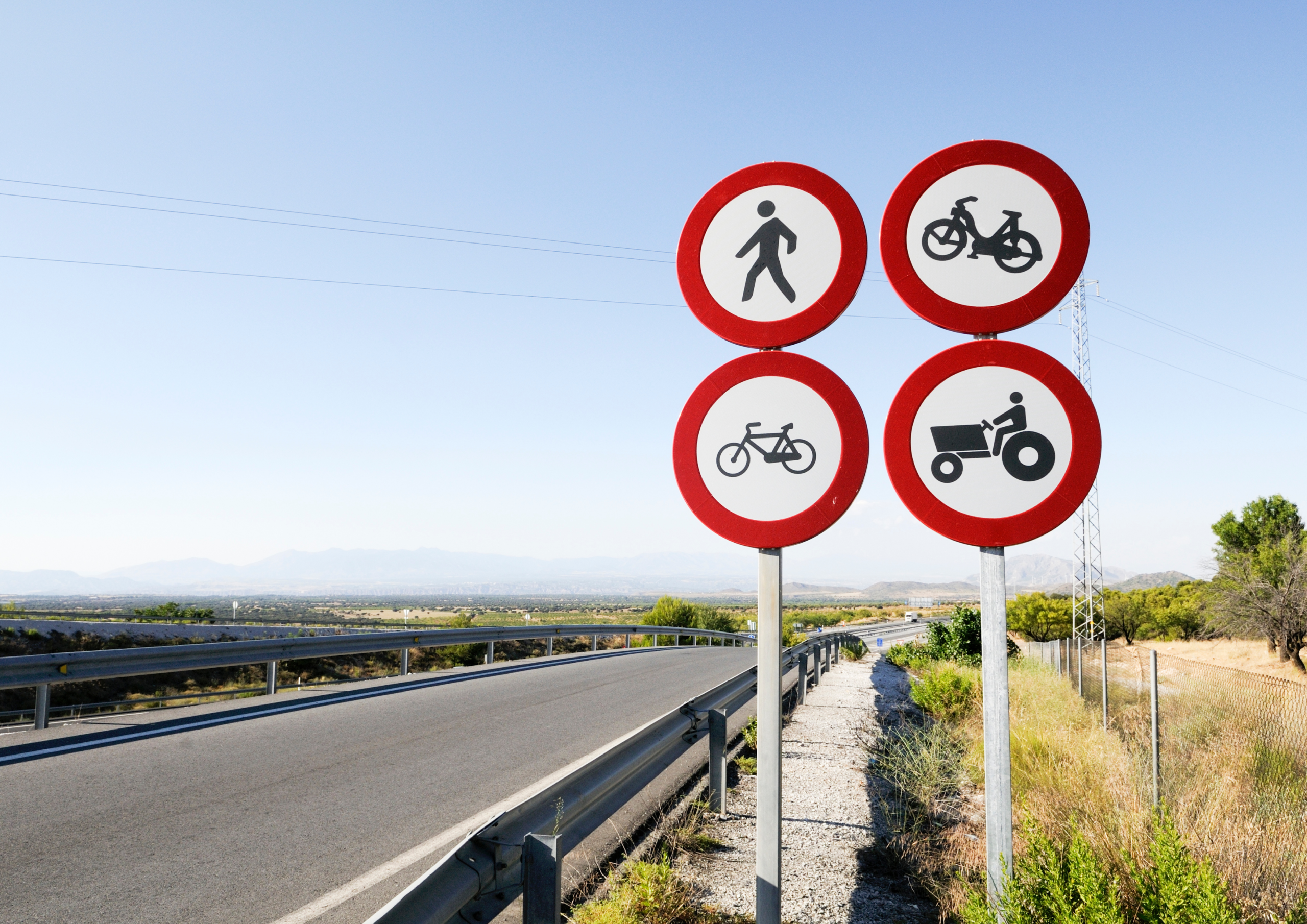 пдр Cluster of four red-and-white no-entry traffic signs: pedestrian, bicycle, motorcycle, and tractor rider on a roadside backdrop with blue sky and desert landscape.
