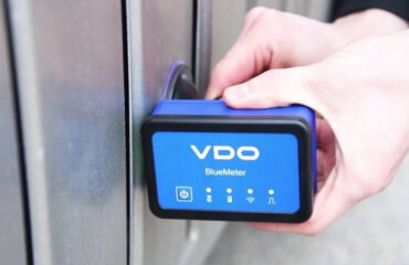 Hand holding a blue VDO BlueMeter device against a metal door lock.