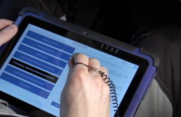 Person using a stylus to interact with a blue-screen tablet in a rugged purple case, selecting menu items.