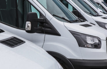Row of white delivery vans parked side by side in a lot, showing their fronts and large mirrors.